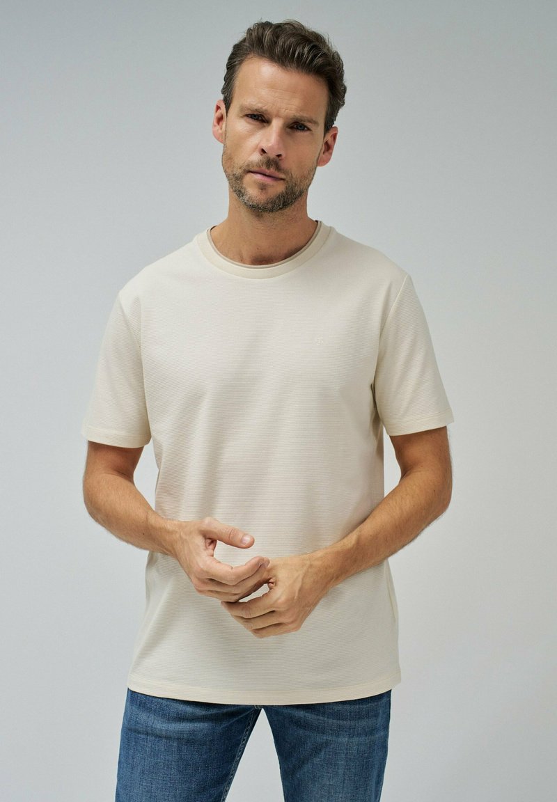 Camiseta beige de manga corta con cuello redondo, tejido texturizado y un sutil logo en el pecho, combinada con jeans azules.