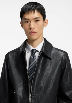 Jeune homme aux cheveux courts noirs portant une veste en cuir noire, une chemise blanche et une cravate rayée, faisant face à un fond blanc.
