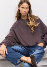 Oversized, langærmet pullover i dæmpet lilla stof med ribstrikkede manchetter, stylet med vidbenede blå denimjeans og mørkebrune mules.