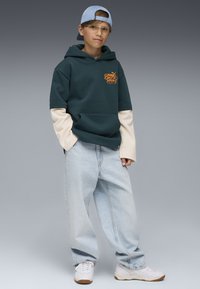 Felpa verde e crema con tasca a marsupio e logo, abbinata a jeans azzurri larghi e sneaker bianche. Un cappellino azzurro chiaro completa il look.