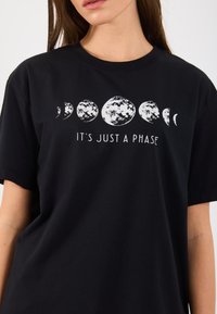 Tricou negru din bumbac, cu un grafic alb reprezentând fazele lunare și textul "E DOAR O FAZĂ" în centru. Design cu mâneci scurte și guler rotund.