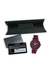 Montre OOZOO avec un boîtier rond bordeaux, bracelet en silicone assorti, cadran bordeaux texturé, aiguilles dorées et logo. Inclus l'emballage et la carte de garantie.