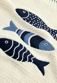 Krämfärgad stickad tröja som har tre broderade fiskar i marinblå och blå mönster, med varierande former och texturer samt kontrasterande konturer.