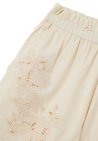 Pantalones beige claros con cintura elástica fruncida y delicados bordados florales con detalles cortados en el lado izquierdo.