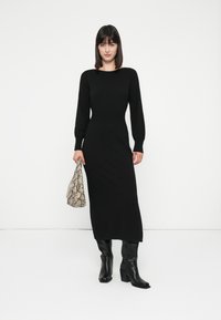 Donna in un lungo abito nero e stivali neri, che tiene in mano una borsa con stampa rettile, in piedi contro uno sfondo bianco semplice.