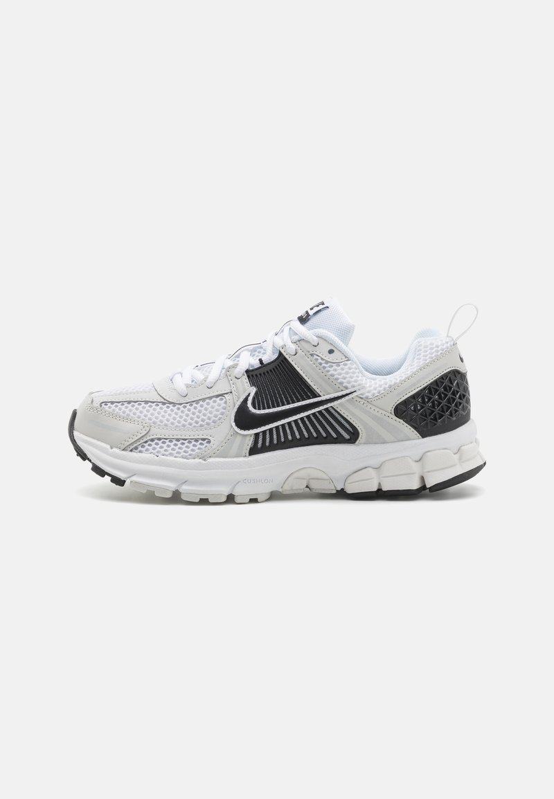 Sapatilha desportiva com parte superior em malha branca e preta, com entressola texturizada e sola de borracha. Inclui atacadores e uma fita de puxar no calcanhar.