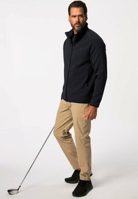 Chaqueta de cremallera azul marino, pantalones chinos caqui, zapatos deportivos negros. Modelo sostiene un palo de golf, mostrando un atuendo relajado pero funcional.