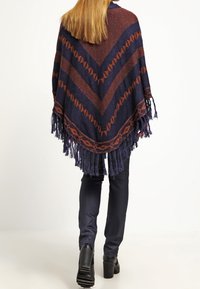 Kvinna med långt hår iklädd en marinblå och rostfärgad mönstrad poncho med fransar, svarta byxor och svarta klackstövlar, som går bort från en vit bakgrund.