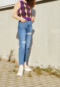 Lila, brun och gräddfärgad argyle-koftväst över en beige långärmad topp, kombinerad med blå jeans med hög midja och slitningar samt vita sneakers.
