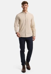 Camisa beige de botones con cuello, de mangas largas; combinada con pantalones navy oscuros y botas marrones con cordones. Tela ligera con textura sutil.