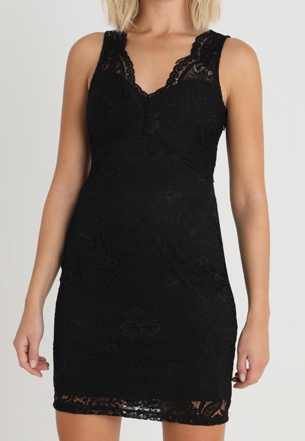 Kvinna som bär en ärmlös svart spetsklänning med V-ringning och blommönster, stående framför en enfärgad ljus bakgrund.