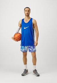 Sinine korvpallivorm, millel on valge Nike'i logo, värviliste mustritega lühikesed püksid ja hallid spordijalatsid. Hoides korvpalli, seisab otse.