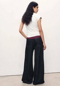 Jean en denim noir évasé avec des coutures contrastantes, assorti d'un t-shirt ajusté blanc avec des manches roulées et une ceinture bordeaux.