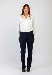 Donna in piedi, che indossa una blusa bianca a maniche lunghe, pantaloni a righe blu navy, scarpe nere e collane sovrapposte su uno sfondo neutro.