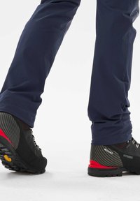 Bottes de randonnée noires et rouges avec des semelles texturées, portées avec un pantalon de plein air bleu marine. Les bottes présentent des lacets métalliques et des accents de design angulaires.