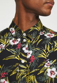 Lyhythihainen kukkapaita mustalla taustalla, jossa on suuria valkoisia ja punaisia kukkia, kirkaan vihreitä lehtiä ja klassinen kaulus.