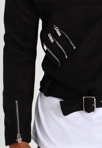 Giacca nera con quattro zip argentate sul petto e una sulla manica, indossata sopra una camicia bianca con una cintura nera con fibbia.