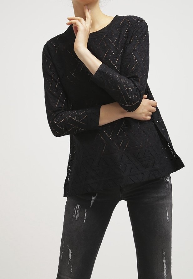 Femme portant un haut noir à manches longues à motifs et un jean gris foncé déchiré, posant avec une main près du cou et l'autre croisée.