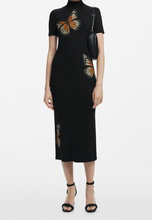 Czarna sukienka midi z krótkim rękawem i pomarańczowymi motywami motyli, czarne szpilki z paskiem na kostce oraz mała czarna torebka na ramię na modelce.