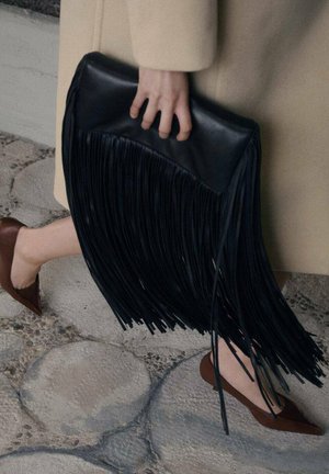 Clutch de cuero negro con flecos largos, forma rectangular, superficie superior lisa, sostenido por una mano. Zapatos planos marrones y un abrigo beige visibles.