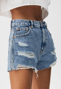 Shorts de denim azul de tiro alto con desgastes y dobladillo deshilachado, usados por una persona con una blusa blanca corta, mostrados desde la cintura hasta la mitad del muslo.