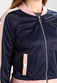 Kvinna som bär en marinblå bomberjacka med ljusrosa detaljer, vita knappar på ärmar och fickor, samt en silverring i dragkedjan.