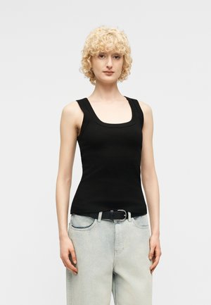 Ung person med krøllet blondt hår iført en sort ærmeløs top og lyseblå jeans med sort bælte, stående foran en hvid baggrund.