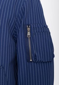 Tessuto blu navy a righe sottili con cerniera metallica verticale e tasca rettangolare sulla manica di un capo d’abbigliamento.