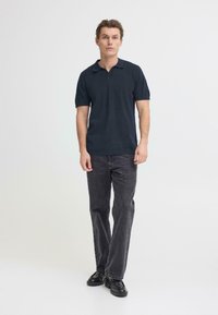 Polo bleu marine à texture douce, manches courtes et col avec revers. Porté avec un jean noir et des chaussures noires, debout sur un fond clair.