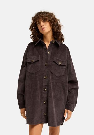 Vrouw met krullend haar die een oversized donkerbruine ribfluwelen blouse met twee borstzakken draagt tegen een effen achtergrond.