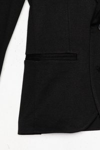 Čierny rukáv saka a dolná časť trupu s klopovým vreckom, zobrazené na textúrovanom bielom pozadí.