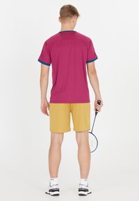 Sportoutfit bestående af en kastanjefarvet t-shirt med korte ærmer og teal detaljer samt gule shorts. Modellen holder et badmintonketcher og står foran en hvid baggrund.