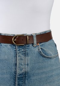 Ceinture en cuir marron avec une boucle en laiton, portée sur un jean en denim bleu clair avec des coutures et des détails de poche visibles.