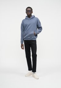 Sweatshirt de algodão azul com bolso frontal, combinado com calças slim-fit pretas e sapatos slip-on bege. Modelo posicionado contra um fundo branco.