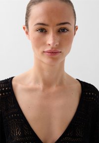 Svart stickad plagg med V-ringning, med intrikata mönster och en halvgenomskinlig textur. Nära bild av en modells överkropp.
