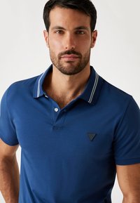 Uomo con capelli corti scuri e barba, che indossa una polo blu con dettagli bianchi sul colletto, su uno sfondo semplice.