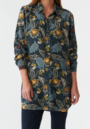 Kvinde iført en langærmet, knappet skjorte med et mørkt blomsterprint i blå, gule og beige toner, kombineret med mørke bukser.