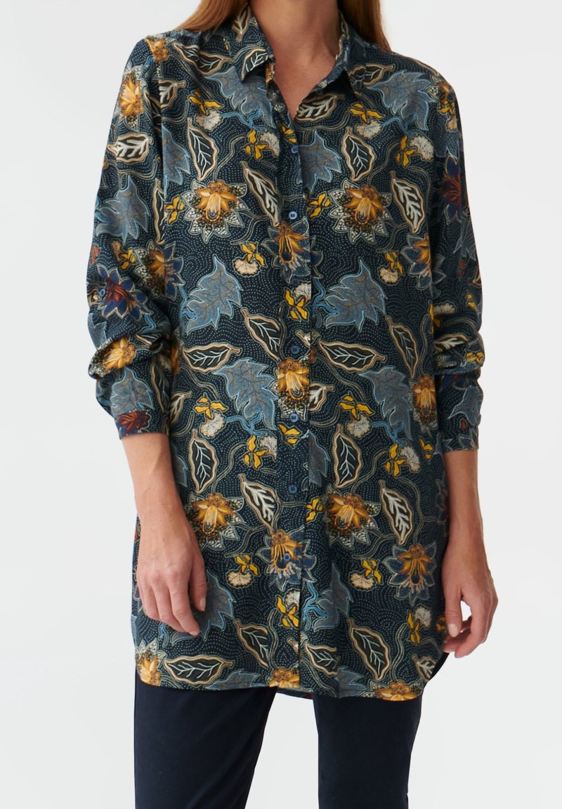 Femme portant une chemise à manches longues boutonnée avec un motif floral foncé dans des tons bleus, jaunes et beiges, associée à un pantalon foncé.