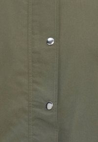 Gros plan sur un tissu vert olive avec une couture verticale et deux boutons-pression argentés brillants.