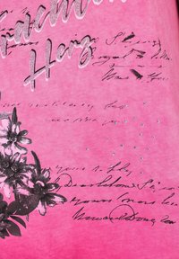 Tela rosa con texto en cursiva negro y estampados florales decorativos. Presenta toques plateados brillantes y pequeñas piedras de strass distribuidas alrededor del texto.