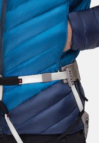 Isolierte Jacke mit einem blauen und marineblauen Steppmuster, ausgestattet mit einem weißen verstellbaren Gürtel und dunklen Akzenten, die die Funktionalität betonen.
