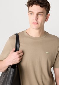 Beige T-shirt med korte ærmer, der har et lille grønt "HUGO" logo. Model med en sort læder skuldertaske.