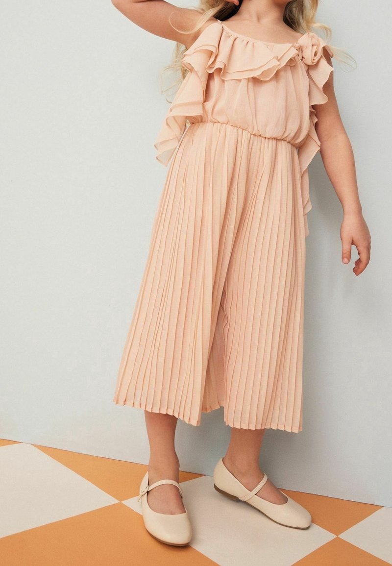 Jeune fille portant un haut rose à volants et un pantalon large plissé avec des chaussures Mary Jane beige, debout sur un sol à carreaux.