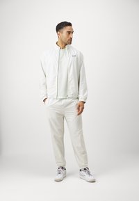 Fato de treino masculino em tom claro, com uma jaqueta com fecho éclair texturizado e calças combinando com punhos elásticos, acompanhados por ténis brancos.