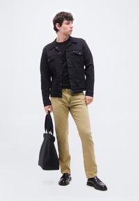 Νέος άνδρας που φοράει μαύρο σακάκι, μαύρο πουκάμισο, μπεζ παντελόνι και μαύρα παπούτσια, κρατώντας μια μαύρη τσάντα tote, στέκεται μπροστά σε απλό ανοιχτό φόντο.