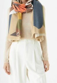 Pull beige et pantalon blanc taille haute portés avec une écharpe multicolore à motif abstrait drapée autour du cou.