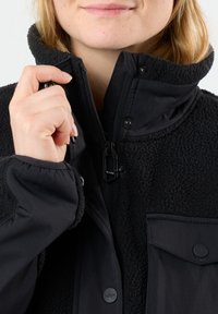 Giacca in pol fleece nera con colletto alto, zip anteriore e bottoni a pressione. Presenta una superficie texturizzata e una tasca sul petto.
