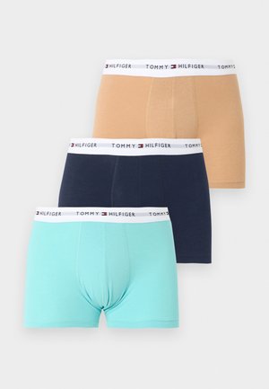 Fiatal, izmos testalkatú férfi, sötétkék Tommy Hilfiger boxeralsót visel, mezítláb áll egy egyszerű fehér háttér előtt.