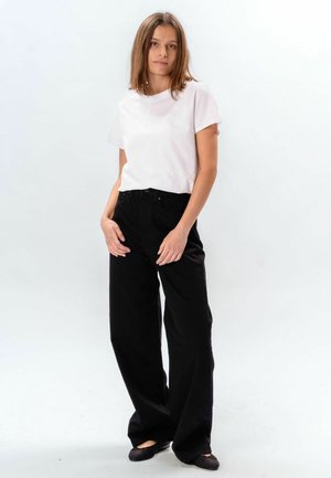 Jeune femme debout portant un t-shirt blanc à manches courtes, un pantalon noir à jambes larges et des ballerines noires, sur un fond clair uni.
