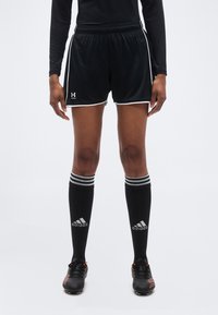 Άτομο που φοράει μαύρο αθλητικό σορτς Under Armour, μαύρες καλτσες Adidas μέχρι το γόνατο με λευκές ρίγες και μαύρα ποδοσφαιρικά παπούτσια με πορτοκαλί λεπτομέρειες.